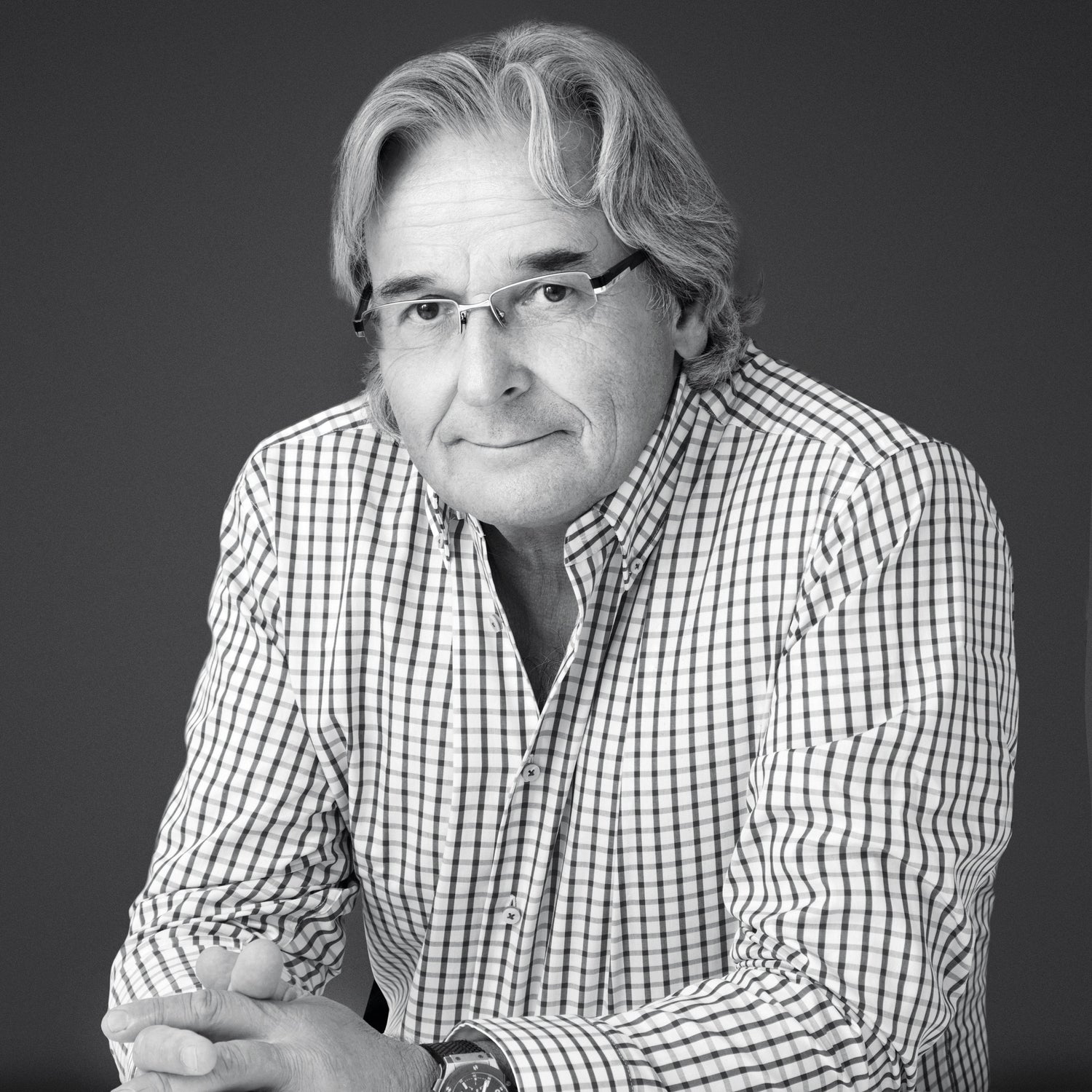 Black-and-white portrait of an older man with glasses and gray hair, wearing a checkered shirt and wristwatch, sitting at a table with his hands folded.