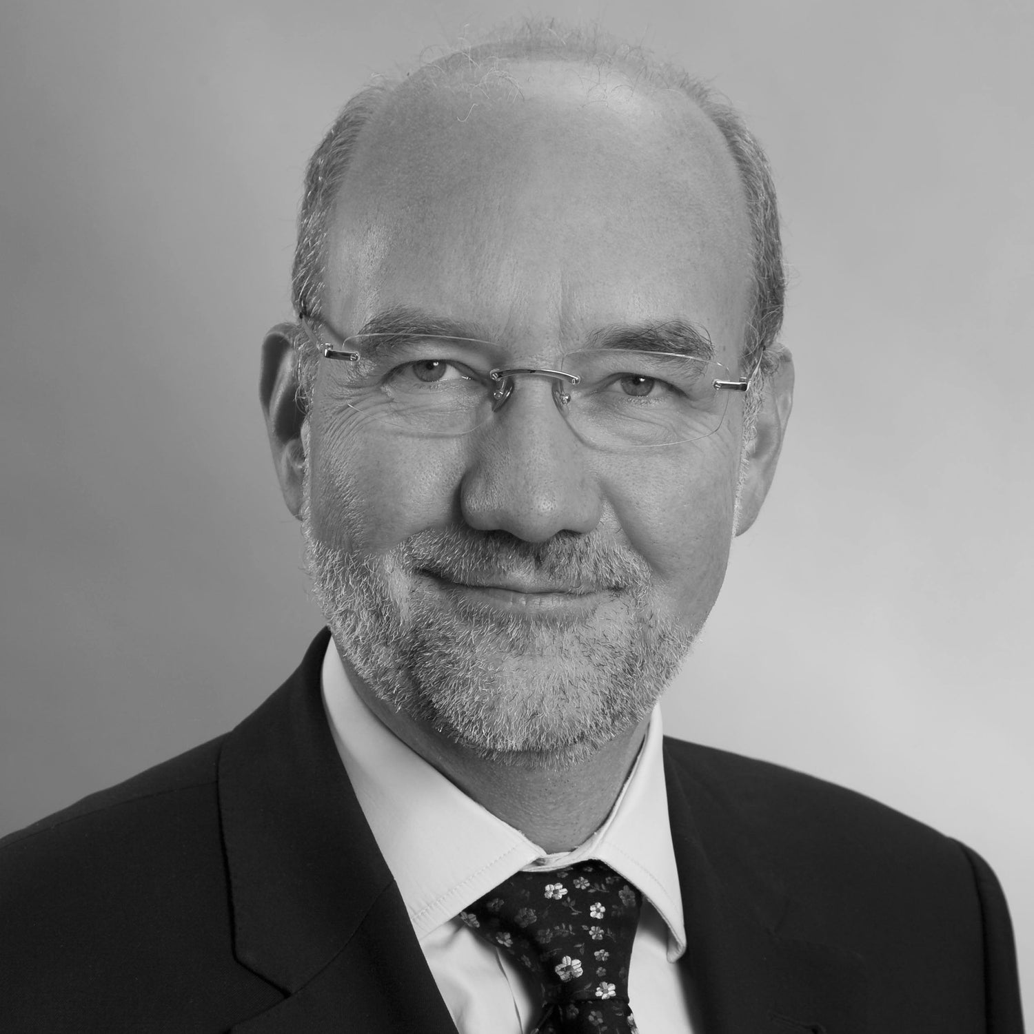 Black-and-white professional portrait of a man with glasses, short beard, and receding hairline, wearing a suit, white shirt, and patterned tie.