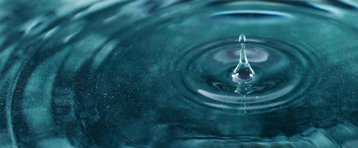 Close-up of a water droplet splashing into a dark teal surface, creating ripples and circular waves.