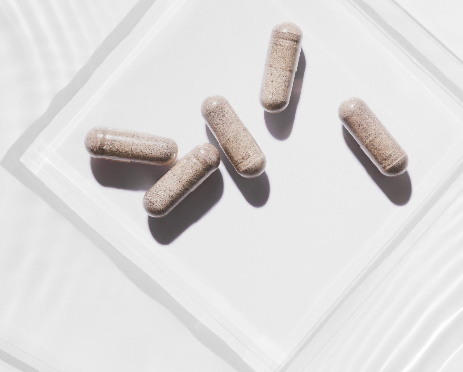 Close-up of five beige supplement capsules arranged on a clear glass surface with soft light and shadows.