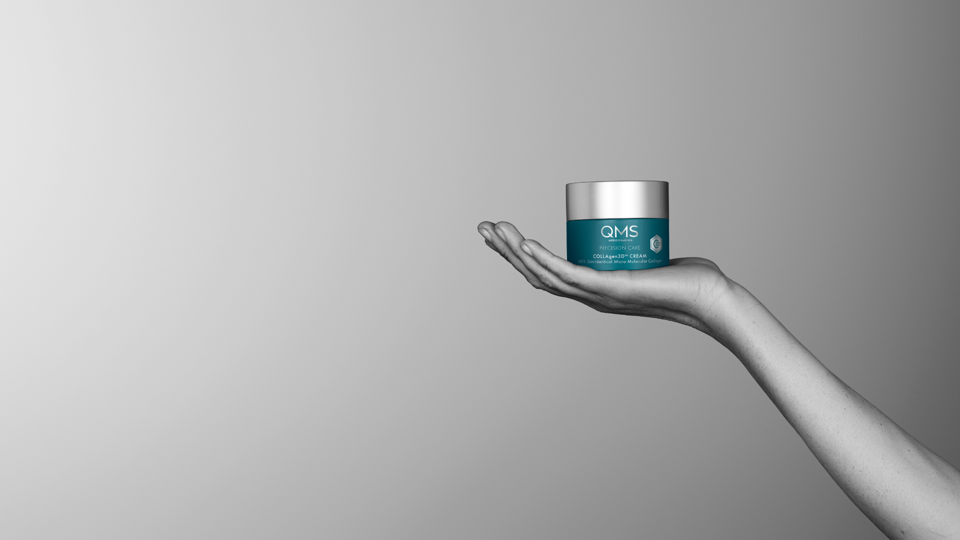 Hand holding a QMS Medicosmetics COLLAgen3D cream jar against a gray background, highlighting the product as the central focus.
