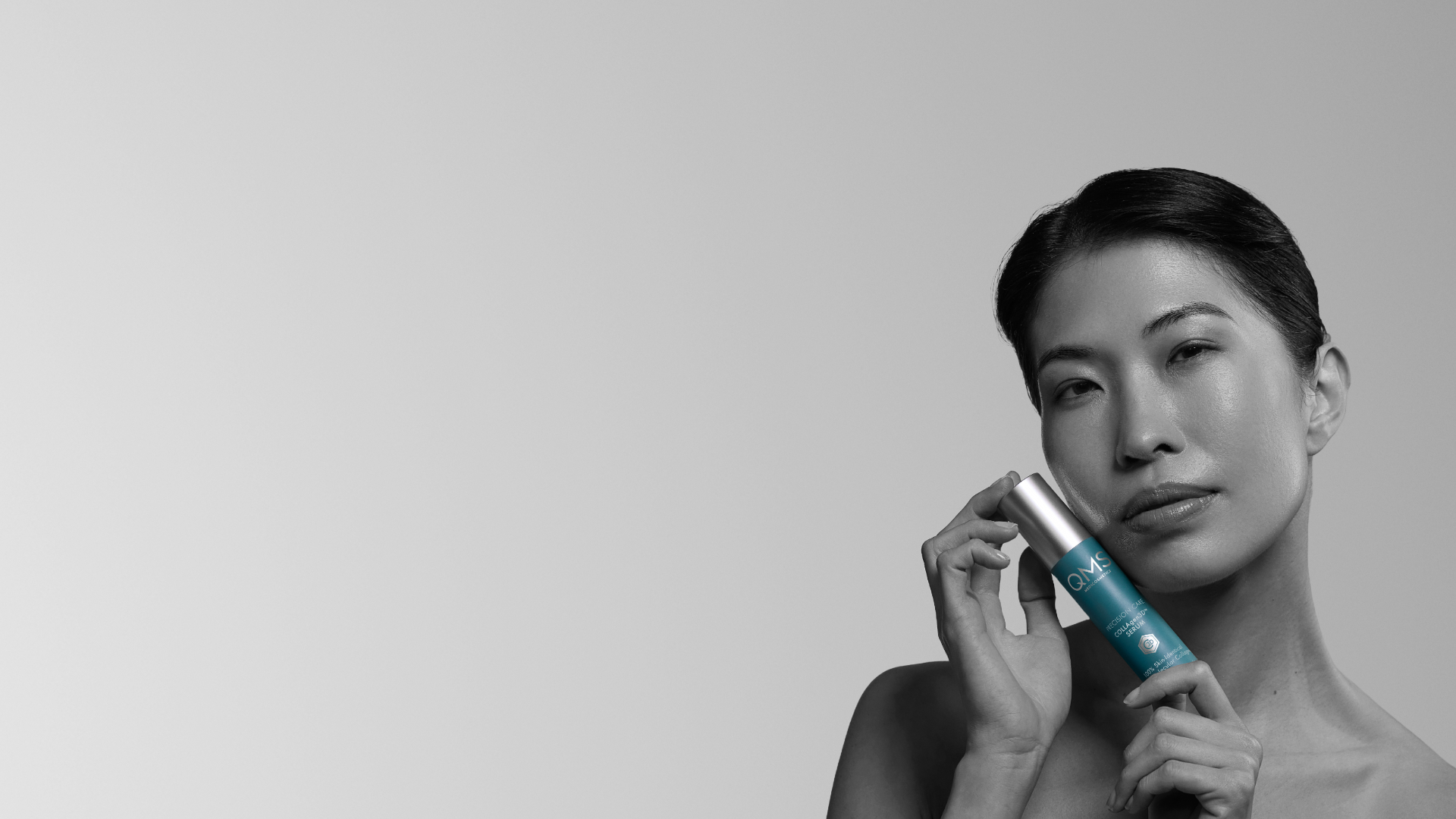 Portrait of a woman holding a QMS Medicosmetics COLLAgen3D light cream bottle beside her face, against a soft gray background.